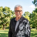 Joe Bonsall, longtime tenor of The Oak Ridge Boys, smiling outdoors in a park wearing a black patterned jacket.